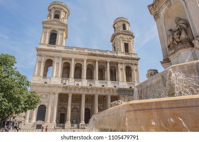 Paris / France - May 18, 2018: Exterior Of The Beautiful And Historic Saint Sulpice Church, Located In The Saint-Germain Des Pres Neighborhood, In Focus In The Distance Beyond The Plaza's Fountain.