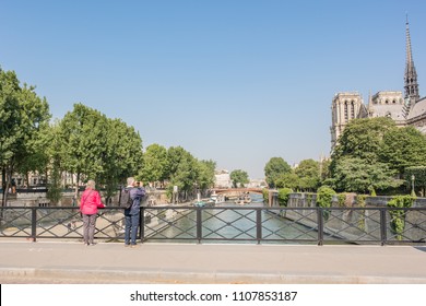 Paris / France - May 19, 2018: An Older Couple Stops To Take A Photo At A Romantic Scene, On A Bridge Overlooking The Seine River And The Notre Dame Cathedral.