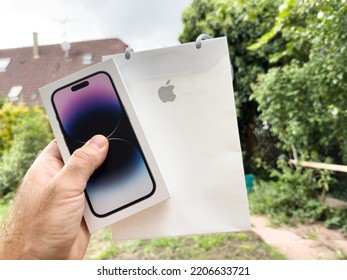 Paris, France - Sep 18, 2022: POV Male Hand Holding New Apple Computers Iphone 14 Pro Cardboard Box With Present Paper Bag With Iconic Logotype - Garden Green Background