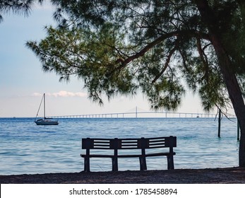 Park bench in silhouette under large evergreen shade tree on barrier island with view of sailboat at anchor some distance from Sunshine Skyway Bridge across Tampa Bay in west central Florida, USA