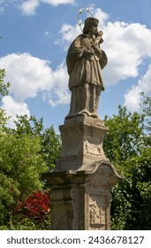 In a park and cemetery of the All Saints church is historical statue of Jan Nepomucky, with a cross in the hnads. Blue sky with white clouds. Dlouhomilov, Moravia, Czech republic.