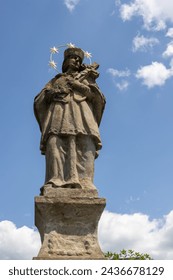 In a park and cemetery of the All Saints church is historical statue of Jan Nepomucky, with a cross in the hnads. Blue sky with white clouds. Dlouhomilov, Moravia, Czech republic.