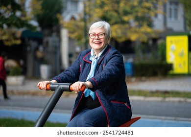 In a park filled with autumn colors, an elderly woman with glasses exercises on outdoor equipment. Her blue clothing and scarf complement the serene seasonal atmosphere - Powered by Shutterstock