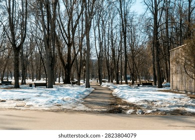 A park pathway meanders through a winter landscape. Bare trees line the path while patches of snow remain on the ground. The sun shines down on the scene, revealing a quiet day.