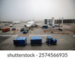 Parked airplane at an airport surrounded by workers and a luggage cart. The aircraft is connected to the boarding bridge but grounded due to heavy fog, preventing flights. Aviation Stock photo 