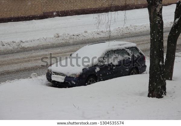 Parked car in the winter in the snowbank on the\
side of the city road - snowfall, cleaning of streets, utilities,\
snow storm