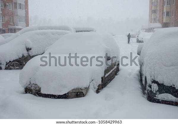parked cars in the yard\
covered with a large layer of snow on the street and snowstorm at\
the end of March