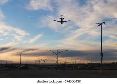 Parking Lot Full Of Cars With Airplane Flying Over At Sunset