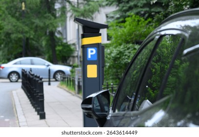 A parking machine is located beside multiple parked cars on a city street surrounded by greenery in bright daylight.