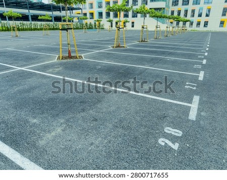 A parking lot shows many empty spaces with numbers marked on the ground. Small trees are placed in the middle of some spaces. A modern building is seen in the background.