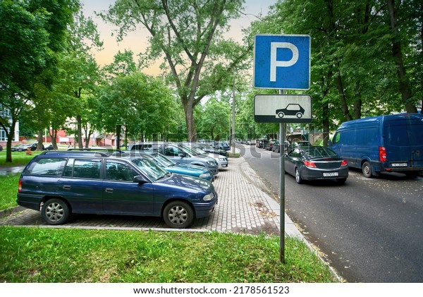 Parking lot sign, cars parked under green trees\
in residential area, Parking zone. Parking problems not enough free\
space. Crowded public parking lot on narrow street. Rows of cars\
parked on roadside