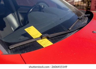 Parking ticket placed under the windshield wiper of a red car. Close-up view of the fine on a dusty windshield in bright daylight. Urban setting, enforcement of traffic regulations.