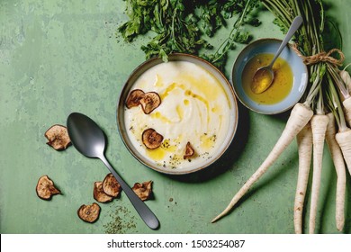 Parsnip cream soup in ceramic bowl with sun dried pears, butter sauce, bundle of fresh parsnip and herbs over green texture background. Flat lay, space