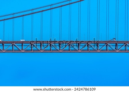 Part of the 25 de Abril suspension bridge with red steel cables that support the structure where cars, vans and trucks circulate to access Lisbon, under a clear blue sky.