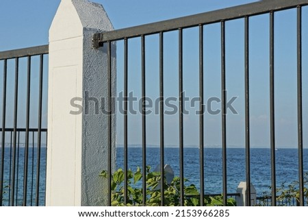 part of a fence wall made of black iron bars and a white concrete pillar against a blue sky and sea