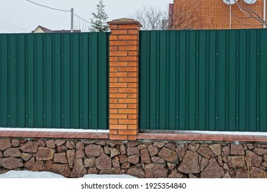 part of a fence wall made of red bricks and green metal on brown stones foundation in white snow on a winter street