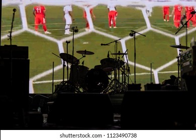 Part Of Football Soccer Game With Players In Red And White And Soccer Net On Goal Showing On A Big Screen Before The Beginning Of The Music Concert, With A Black Silhouette Of A Drum Set In Front