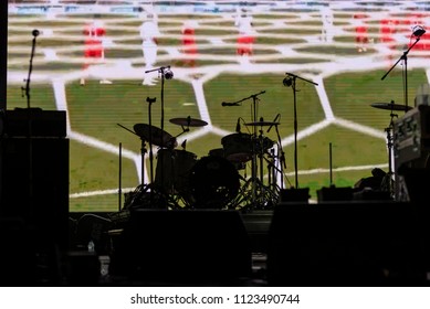 Part Of Football Soccer Game With Players In Red And White And Soccer Net On Goal Showing On A Big Screen Before The Beginning Of The Music Concert, With A Black Silhouette Of A Drum Set In Front