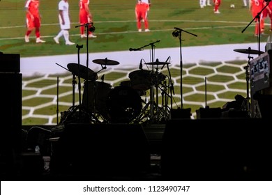 Part Of Football Soccer Game With Players In Red And White And Soccer Net On Goal Showing On A Big Screen Before The Beginning Of The Music Concert, With A Black Silhouette Of A Drum Set In Front