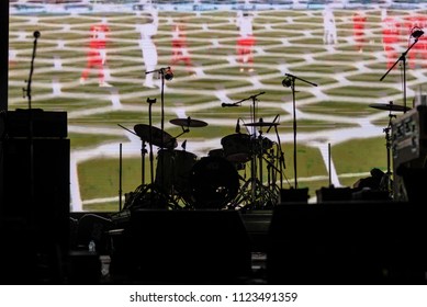 Part Of Football Soccer Game With Players In Red And White And Soccer Net On Goal Showing On A Big Screen Before The Beginning Of The Music Concert, With A Black Silhouette Of A Drum Set In Front