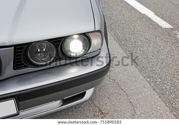 Part of the gray car on the background\
of asphalt. The car\'s headlights are shining. Lights burn. Car\
headlights. Luxury Headlights. Gray car. Car\
details
