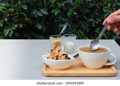 Part Of Hand Holding Spoon To Stir Coffee And There Are Cookies, Sugar And Milk Together On Wooden Tray. Garden Atmosphere.