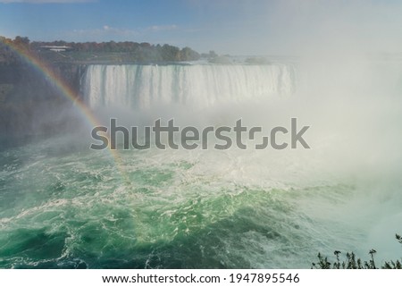 A part of Horseshoe Falls with a tall cloud of water splashes, rainbow, and several people walking on the embankment. View from the opposite Canadian shore