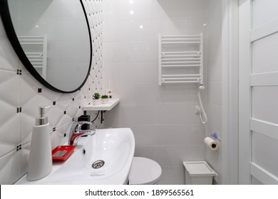 A Part Of A Modern Small Bathroom In A Newly Furnished Studio Apartment In An Old House. Ceramic Tiled Walls, Towel Radiator On White Wall, Sink, Mirror, Shelf With Flowers, Door, Toilet, Laundry Bag