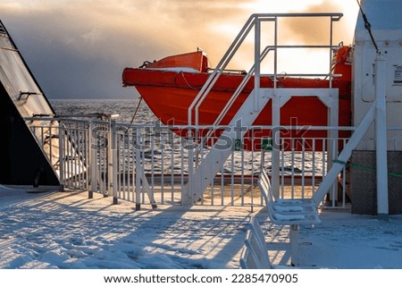 Part of orange lifeboat on snow and ice-covered ferry deck, dramatic and sunny sky in sunset. From Moskenes to Bodø, Lofoten, Norway.