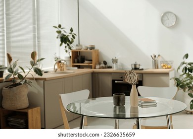 Part of spacious kitchen with round glass table with vases and notepads standing in the center against green domestic plants and counter