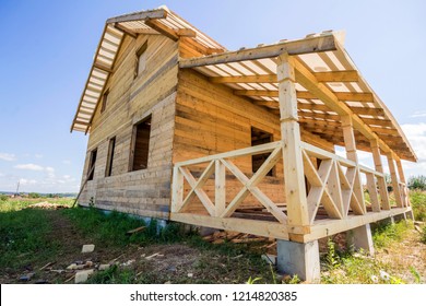 Part of unfinished wooden ecological traditional cottage of natural lumber materials with steep roof frame and attached terrace with decorative railing under construction in green neighborhood.