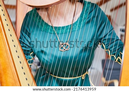 Part of a woman, wearing a keltic knot and a traditional green dress visible through harp strings