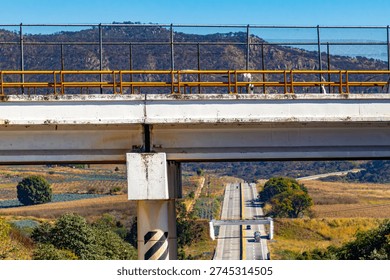 Partial aerial view of elevated vehicular bridge with yellow fence and cyclone mesh, two-way road between farmland below, mountain against blue sky in background, sunny day in Jalisco, Mexico