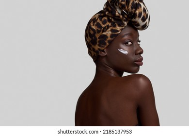 Partial Back Of Elegant Black Girl With Cosmetic Cream On Face. Beautiful Young Slim Woman Wearing Traditional African Turban. Body And Skin Care. White Background. Studio Shoot. Copy Space