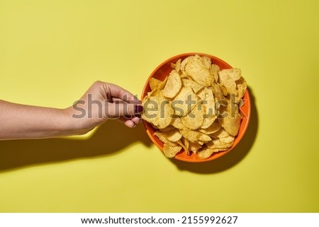 Partial female hand taking tasty and appetizing potato chip from bowl. Unhealthy eating and fast food. Crunchy snack for leisure. Isolated on yellow background. Studio shoot. Copy space. Top view