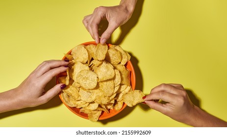 Partial Female Hands Taking Tasty And Appetizing Potato Chips From Bowl. Unhealthy Eating And Fast Food. Crunchy Snack For Leisure. Isolated On Yellow Background. Studio Shoot. Copy Space. Top View