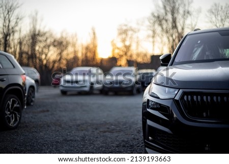 Partial front view of new black modern family SUV. Visible front LED light and part of gloss black grille and bumper. Out-of-focus cars and trees in the background. Sunset light. Parking on gravel.