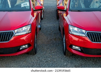 Partial front view of two new red passenger cars. Visible headlamps, foglamps and part of grilles, bonnets and bumpers. Sunset light. Parking on gravel. Symmetrical composition. Paper with number.