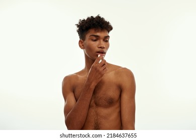 Partial image of black guy moisturizing his lips with hygienic lipstick. Young brunette curly man with naked torso. Body and skin care. Isolated on white background. Studio shoot. Copy space