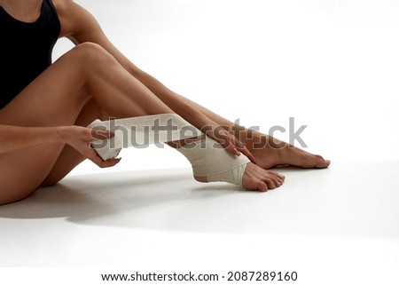 Partial image of female ballet dancer putting elastic bandage on her leg. Choreography concept. Woman barefoot and wearing leotard. Girl isolated on white background. Studio shoot. Copy space