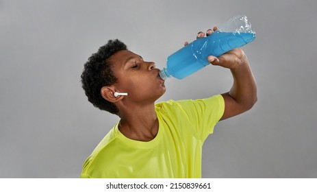 Partial Image Of Sportive Black Boy With Closed Eyes Drinking Blue Sport Drink From Bottle. Healthy Lifestyle. Male Child Wearing Wireless Earphones And Yellow T-shirt. Grey Background In Studio.