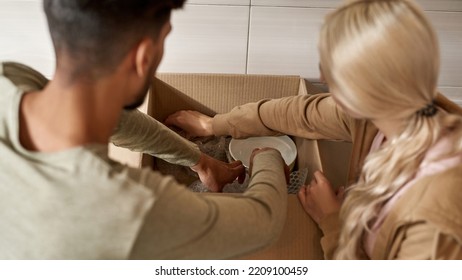 Partial Image Of Young Couple Packing Cooking Utensils In Air Bubble Film In Cardboard Box At Kitchen. Blonde Girl With Tattoo And Bearded Man Moving To New Apartment. Concept Of Home Relocation