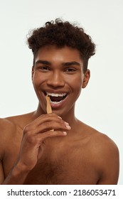 Partial Of Pleased Black Guy Brushing His Teeth With Toothpaste. Young Brunette Curly Man With Naked Torso Looking At Camera. Dental Care And Hygiene. Isolated On White Background. Studio Shoot