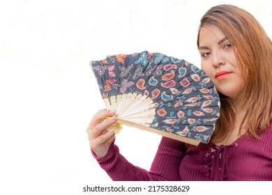 Partial Portrait Of A Young Mexican Woman With An Open Fan, Serious Expression, Against A White Studio Background, Long Straight Brown Hair, Fashion Eyebrows, Natural Makeup, Casual Violet Blouse