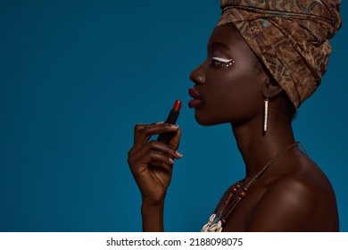 Partial Side View Of Stylish Black Girl Painting Lips With Lipstick. Attractive Young Woman Wearing Traditional African Outfit And Accessories. Female Beauty. Blue Background. Studio Shoot. Copy