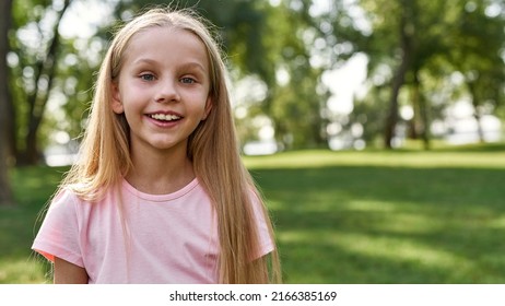 Partial Of Smiling Caucasian Girl Looking At Camera On Green Meadow In Blurred Park. Cute Blonde Female Child Of Generation Alpha Wearing T-shirt. Concept Of Childhood Lifestyle. Sunny Summer Day