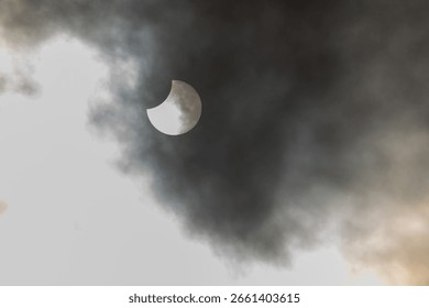 A partial solar eclipse occurs as the moon obscures part of the sun, surrounded by dynamic cloud formations. The event takes place in the daytime, providing a stunning visual experience. France