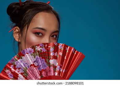 Partial of stylish asian girl covering half face with traditional handheld fan. Attractive young brunette woman looking at camera. Female beauty. Isolated on blue background. Studio shoot. Copy - Shutterstock ID 2188098101