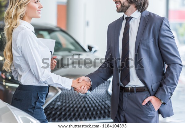 partial view of auto salon seller and\
smiling businessman shaking hands at dealership\
salon