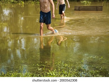 Partial View Of Bodies Of Young Man And Teenage Boy Walking Through Water Of Flooded Midwestern Park; Park Bench Immersed In Water Behind Them